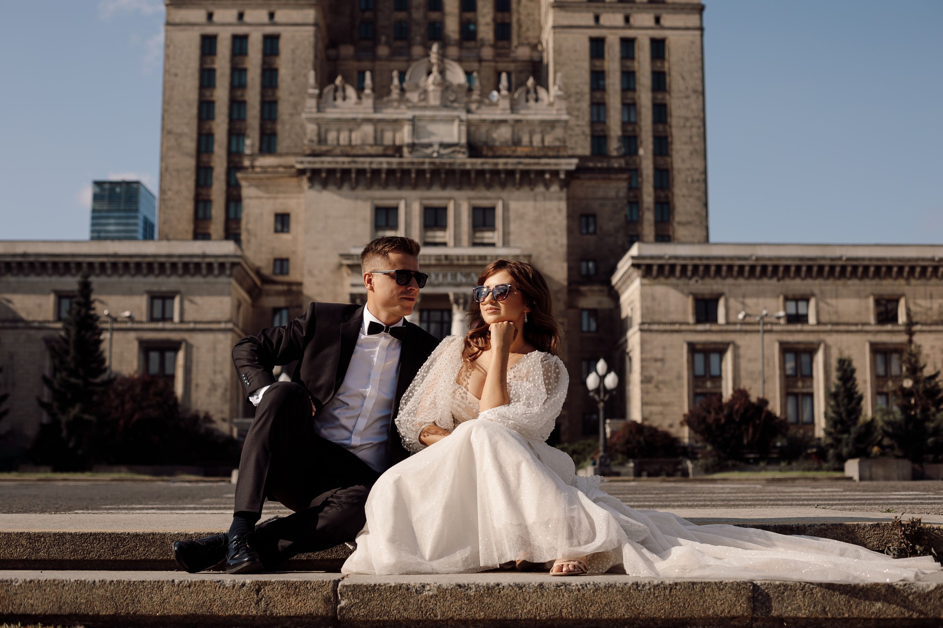 una joven pareja enamorada, la novia y el novio con vestido de novia están sentados en los escalones contra el telón de fondo del paisaje urbano en gafas de sol felices juntos día soleado